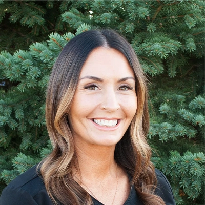 Chiropractic Assistant Josee Smiling Outdoors In Front Of Pine Tree