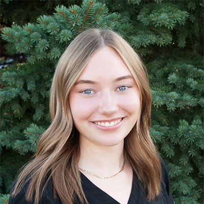 Chiropractic Assistant Milee Smiling Outdoors In Front Of Pine Tree